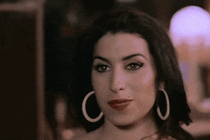 Celebrity gif. Amy Winehouse smiles flirtatiously as she softly waves. 