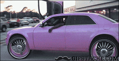 Video gif. Pink modified car with huge tires and rims races through a parking lot with a door up, showing off, as a police car chases it.