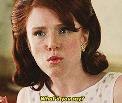 TV gif. A young woman seems to be in the middle of eating something as she speaks to us with irritated surprise. Her red, shoulder-length hair is cut in a retro 60s style. Text, "What'd you say?"