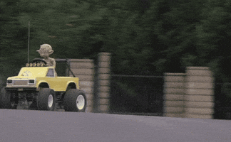 Video gif. An alien in an automatic mini car racing down a neighborhood street, drifting on the turn. 
