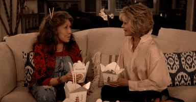 TV gif. Lily Tomlin as Frankie and Jane Fonda as Grace from Grace and Frankie sit facing each other on a white couch. They toast with two open cartons of Chinese food as if saying "cheers!"