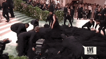 Met Gala 2024 gif. Cardi B appears in her strapless Windowsin black gown gesturing to one of seven attendants assisting with her giant trumpet skirt that flares out about 10 feet in all directions, nearly filling the red carpet area. The attendants are fluffing the billowing fabric so it lays in a perfect circle.