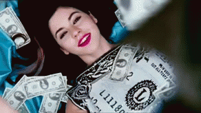 Video gif. A woman lays on the floor wearing a dollar bill dress. Cash money is poured over top of her like itâ€™s raining dollar bills. She smiles happily and holds her hands out. 