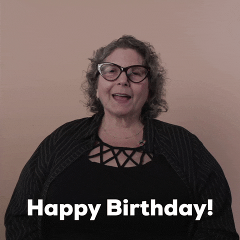 Reaction gif. A Disabled white woman with kinky curly gray hair and big wine-colored cat-eye glasses says happily and directly, "Happy birthday!"