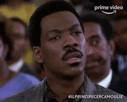 Movie gif. Eddie Murphy as Prince Akeem in Coming to America sits in a crowd of people. He looks straight ahead and looks away confused. He blinks quickly as if processing what he’s just heard. 
