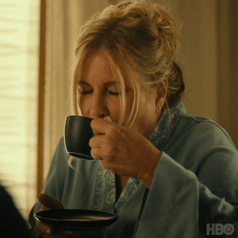 TV gif. Jennifer Coolidge as Tanya on The White Lotus. She is holding a coffee cup and a saucer and she takes a much needed sip from the cup with her eyes closed. She opens her eyes slightly and nods her approval before going in for another sip.