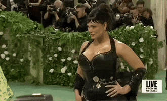 Met Gala 2024 gif. Ashley Graham wearing a custom Ludovic de Saint Sernin gown with black leather bodice, offers moody poses to the paparazzi as someone runs through frame behind her.