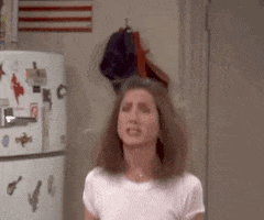 Friends gif. Jennifer Aniston as Rachel stands in a room with a magnet covered refrigerator. She jumps and waves her hands in the air, smiling broadly and covering her mouth with excited hands. 