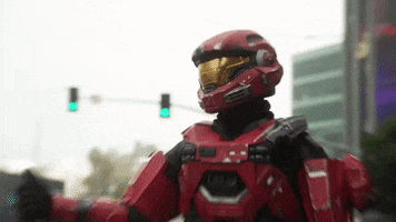 Video game gif. Red spartan from Halo stands on the side of a city street with his thumb out to hitchhike as cars drive past.