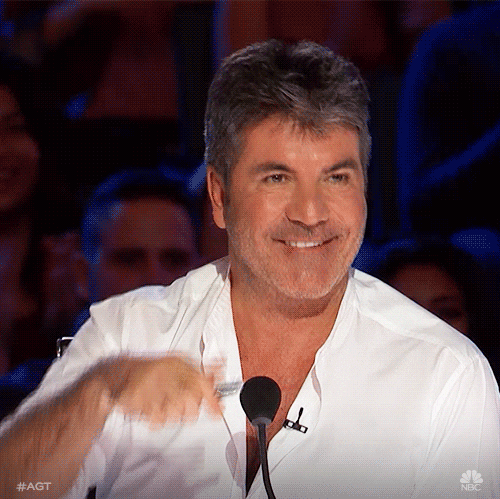Reality TV gif. Simon Cowell on America’s Got Talent sits at the judges desk and looks up at the stage with a wide earnest smile. He gives a big thumbs up as the crowd behind him cheers and claps.