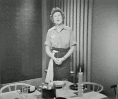 TV gif. In black and white, Julia Child folds her hands in front of a table set for dinner and says, “Bon Appetit.”