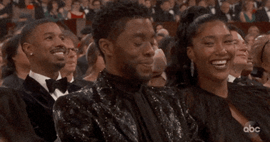 Video gif. Chadwick Boseman sits in the audience at the Oscars looking down and laughing with a shy or flattered expression as his date taps him on the arm and gives him a knowing smile. 