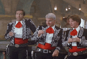 Video gif. Three men dressed in matching black mariachi outfits with bright red cummerbunds and frilly neckties all take a shot. At first, they show no reaction, but then suddenly spaz out for a moment before reeling themselves back in.