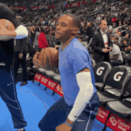 Sports gif. Video of NBA player Russell Westbrook working it out in an energetic dance by himself wearing casual clothes, bending his knees and popping his hips back and forth with swagger as he holds a basketball in one hand with a stadium of fans behind him, mouth curled in a straight line like he's focusing on the moves. 
