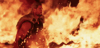 Movie gif. Chris Hemsworth as Thor saunters through blazing flames as smoke wafts around him.