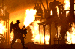 Video game gif. Scene from Team Fortress 2 shows a set of buildings engulfed in flames, while a person in the foreground hoists up a flamethrower toward it.