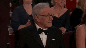 Oscars 2024 GIF. Steven Spielberg turns to the camera and uses his right hand to point at himself, shaking his head sadly. 
