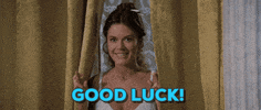 Video gif. Brunette woman stands between yellow drapes and grins, holding up both hands with fingers crossed. Text, "good luck!"