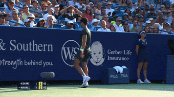 Sports gif. Clip of Carlos Alcaraz on a competition tennis court walking away from us towards the back wall of the court. He does two push ups like he's showing how strong he is, then turns towards us and smiles. 