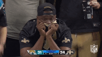 Video gif. Stressed or disappointed New Orleans Saints fan covers his mouth with the collar of his shirt. He's the only one still seated as all the other fans around him stand up and gesture at the field.