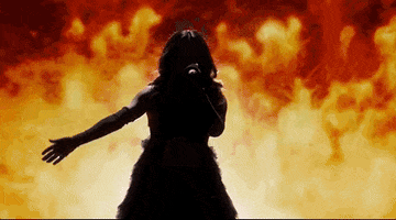 Oscars 2024 GIF. Becky G performing The Fire Inside from Flamin' Hot. She's shrouded in shadows and the spotlight suddenly beams on her. She sashays into the center, waving a hand sensually as she sings into the microphone. She wears a black dress with a cut out on her torso, long black gloves, and flames shoot up behind her. 