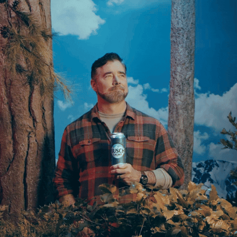 Sponsored gif. Gerald Downey wearing a red and black plaid shirt holding a can of Busch Light beer at his chest. He takes a long, deep breath as if he's taking in the beautiful mountain scenery around him, exhaling with a smile and nod, looking around with wonder and appreciation. Text, "TGIF."