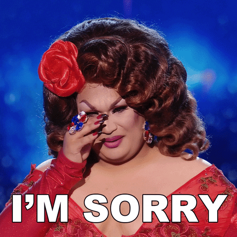 TV gif. In a scene from Queen of the Universe, wearing a red dress and a large red flower ornament in their hair, a smiling Ada Vox puts their hands together in gratitude. Text, "I'm sorry."