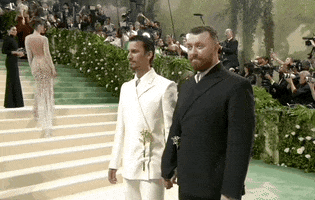 Met Gala 2024 gif. Sam Smith and designer Christian Cowan stand awkwardly, allowing for pictures, wearing matching suits, Sam in black, Christian in ivory.