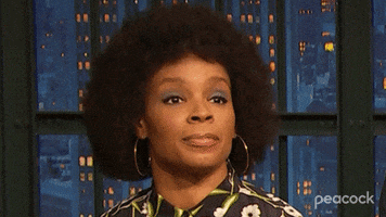 TV gif. Closeup of Amber Ruffin as she tilts her head to the side and shrugs with a frown on the set of her eponymous show.
