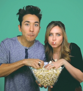 Video gif. A young man and woman scarf popcorn while staring intently at us, eager to see what happens next.
