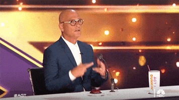 Reality TV gif. On the set of AGT, Howie Mandel gives us a tense look as he crosses his fingers on both hands.