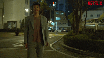 TV gif. Lee Jung-jae as Seong Gi-hun in Squid Game stands on a street, waving and smiling sheepishly.