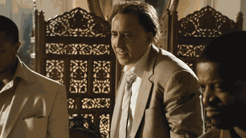 Celebrity gif. Nicolas Cage stands amongst other men very still with an awkward toothy smile and blank stare. We zoom in on him and he barely moves like he’s a statue.