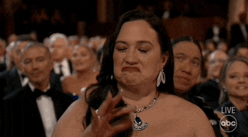 Oscars 2024 GIF. Lily Gladstone, seated at the Oscars, shrugs and nods, gesturing “so-so,” half-confirming.