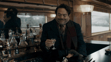 Movie gif. Dan Fogler as Jacob in Fantastic Beasts: Secrets of Dumbledore. He's at the bar inside of a train and he holds a little cocktail glasses while spluttering with laughter.