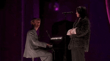 Oscars 2024 GIF. Billie Eilish and Finneas performing "What Was I Made For" on stage at the Oscars. They finish the song and Eilish holds her hands together in front of her and gives a slight bow of appreciation at the standing ovation. Finneas looks at her with pride and claps strongly for her as well.