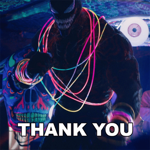 Movie gif. Tom Hardy as the character Venom wearing glow-stick necklaces on stage, pointing and speaking into a microphone, "thank you."