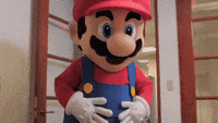 Video game gif. A life sized Mario walks into a room and waves at us.