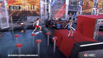 Reality TV gif. Two women race each other up a wall on American Ninja Warrior. The girl in the blue shirt pushes the button first and the girl in the white shirt collapses. The blue shirt woman collapses on top of the other girl.