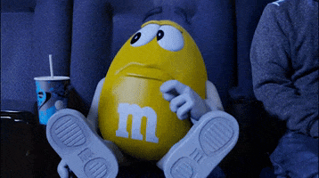 Celebrity gif. The yellow M&M sits at the movies, a soda in his cup holder. He looks down worried and holds his stomach--it's grumbling. Text, "Feed me"