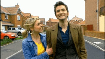 TV gif. Walking down a residential street, Billie Piper as Rose smiles and pulls on the arm of David Tennant as Doctor Who, who smiles as they walk towards us.