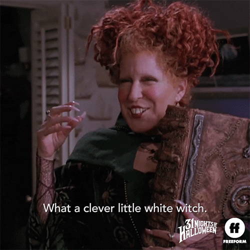 Movie gif. Bette Midler as Winifred in Hocus Pocus. She carries a spell book and gives us a witchy grin and waves softly with her hand while saying sarcastically, "What a clever little white witch."