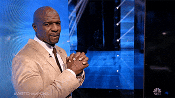 Reality TV gif. Terry Crews from AGT stands backstage in a beige suit. As he glances at us, he sighs apprehensively and crosses his fingers with both hands.