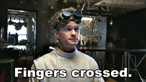 Movie gif. Neil Patrick Harris as Billy in "Dr. Horrible's Sing-Along Blog" looks at us and holds up his hand with two fingers crossed, and says, "Fingers crossed," which appears as text.