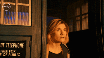 TV gif. Jodie Whittaker as The Doctor in Doctor Who steps out of the Tardis into golden light and looks around.