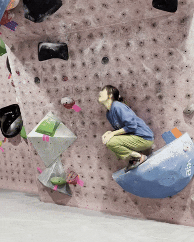 Video gif. Woman in a purple sweater and green pants hops down like a rabbit from a wide purple rock mounted on a rock climbing wall. She lands on a mat in a crouched position. 