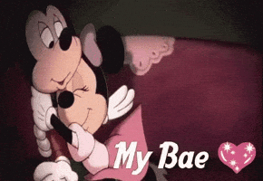 Disney gif. Minnie Mouse tightly hugs Mickey Mouse on a couch. Mickey melts into the hug and embraces her back. They nuzzle their heads closer to each other. The text says, “My Bae,” with a sparkly heart next to it. 