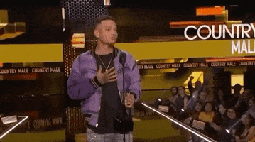 Celebrity gif. Kane Brown at the 2018 American Music Awards steps back from the mic and taps his chest, holding an award with his other hand.