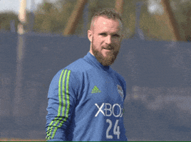 Sports gif. Stefan Frei looks up as if happy to see us and smiles as he waves. 