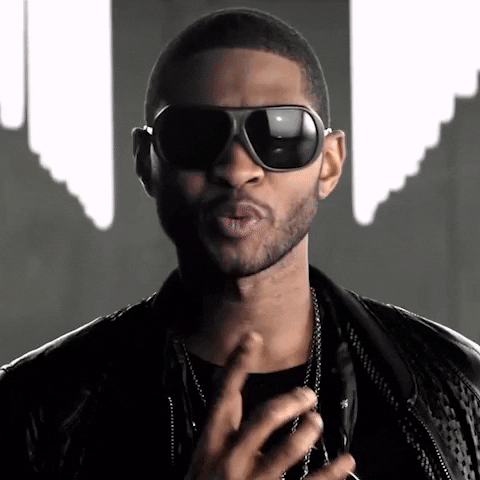 Celebrity gif. Usher wears sunglasses and a leather jacket as he sassily points at us and sings "Somebody to love."